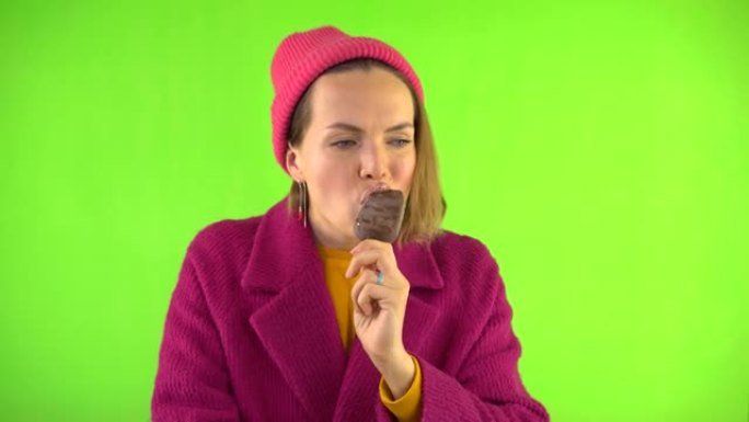女人在巧克力釉的棍子上咬冰淇淋，然后冷冻，她变冷了。女人穿着针织帽，红色外套在工作室的绿色背景