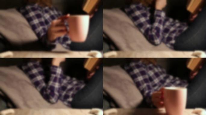 模糊运动。女人喝一杯热咖啡或茶，在沙发上看书。自我隔离时间。检疫时期的娱乐。