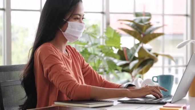 亚洲女性内容创作者戴着口罩，在家里工作的笔记本电脑上的社交媒体平台上打字内容。使用技术的新常态生活方