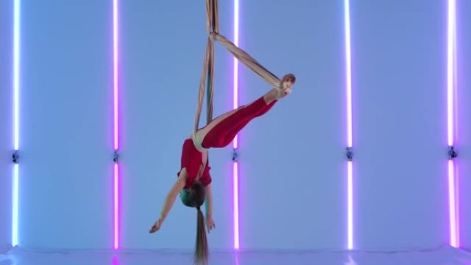 空中体操运动员在工作室里用丝绸表演空中杂技舞蹈。一个穿着红色紧身衣的年轻女孩在明亮的霓虹灯背景下展示