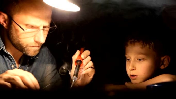父亲在台灯下焊接东西，儿子看着他并谈论它。家庭父母和孩子关系概念镜头。
