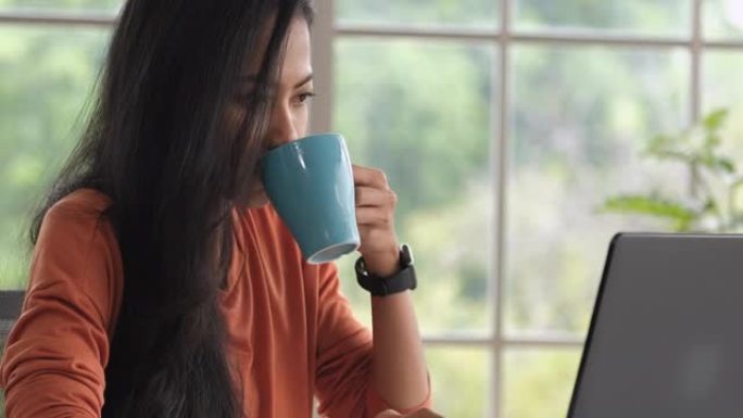 亚洲女性在家工作，一边用笔记本电脑在线工作一边喝咖啡。新的正常生活方式