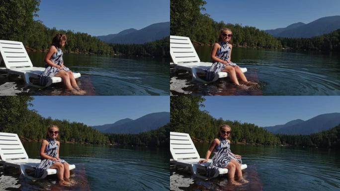 小女孩戴着太阳镜和波西米亚丝绸披肩躺在日光浴床上。儿童在洪水木水下码头上休息。湖面上铺满了水。
