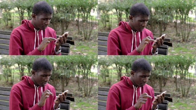 年轻的美国非洲裔小伙子在公园里用智能手机打字