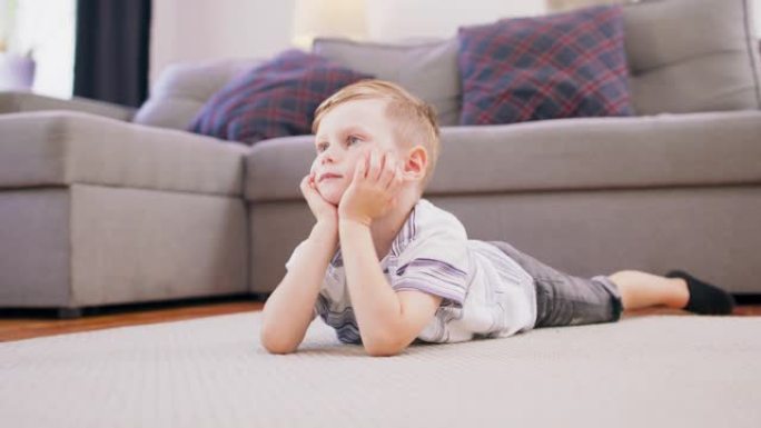 可爱的金发小男孩躺在地板上盯着电视上他最喜欢的动画片