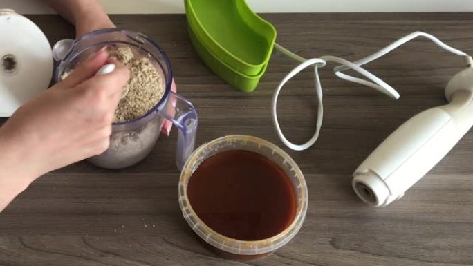 一名妇女取下搅拌机的盖子，用勺子切碎的花生和种子混合，制成halva。容器旁边是蜂蜜，可以添加到混合