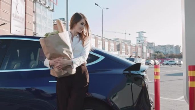 美丽的女商人站在她的电动汽车旁，带着杂货。电动汽车充电。环保运输完成自动充电。附近的高速充电站