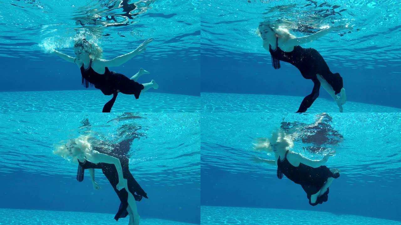 一个年轻的金发女孩穿着黑色连衣裙在水面附近的水下游泳，在水中徘徊，双臂伸出。慢动作。从池底看。