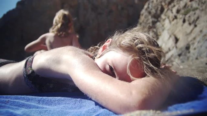 孩子用蓝色毛巾在沙滩上晒日光浴，对着镜头挥手微笑