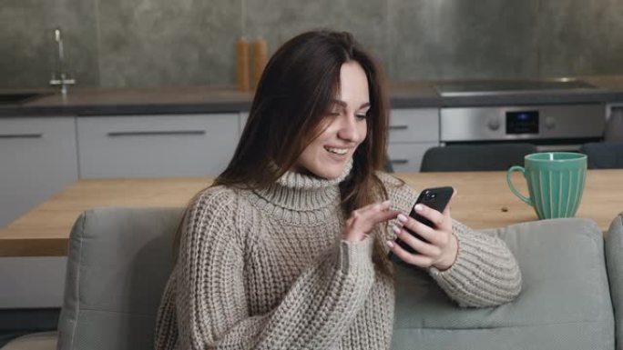 25岁的年轻白人妇女坐在家里的沙发上，穿着一件温暖的毛衣，在现代公寓的智能手机上快速浏览互联网页面。