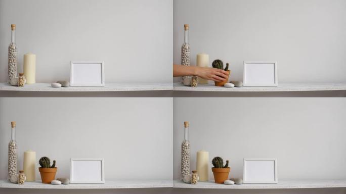 带相框模型的现代房间装饰。搁板靠着白色墙壁，配有装饰蜡烛、玻璃和岩石。手放下盆栽仙人掌植物。
