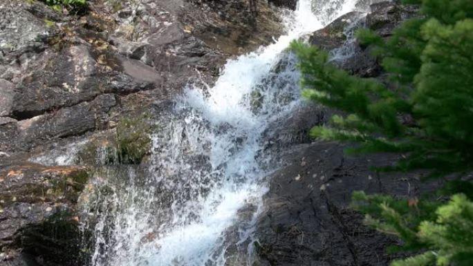 瀑布沿着石墙层叠。阳光照在悬崖和山溪上。前景是云杉分支。石头上覆盖着苔藓。