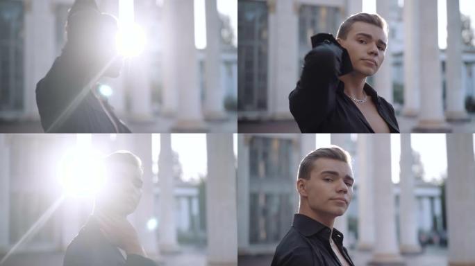 侧视图头部拍摄的自信年轻时尚男子调整头发在阳光和转向相机。英俊的白人芭蕾舞者站在阳光下的城市的肖像