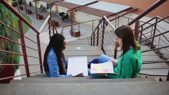 两名年轻的大学女性在彼此聊天时坐在木制楼梯上学习