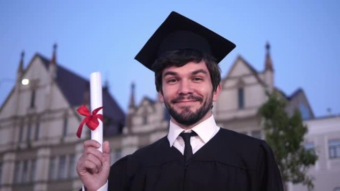 高加索人快乐的年轻毕业男子对着镜头摆姿势，在大学前展示他的文凭