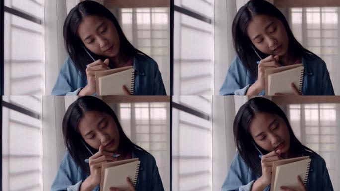 年轻的亚洲女商人站在窗户旁打电话，并在家庭办公室做笔记想法规划策略。