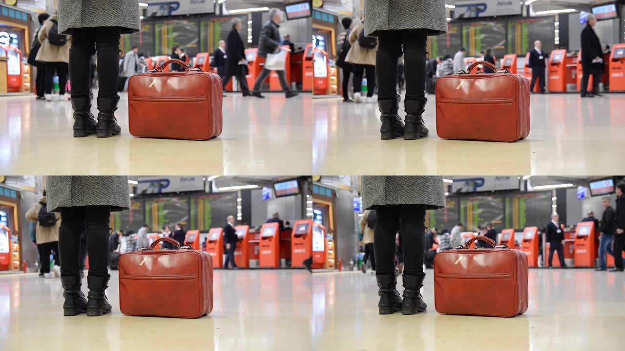 在机场，穿着靴子和红色手提袋的女人，还有一些散焦的人。我们只看到腿和红色袋子。