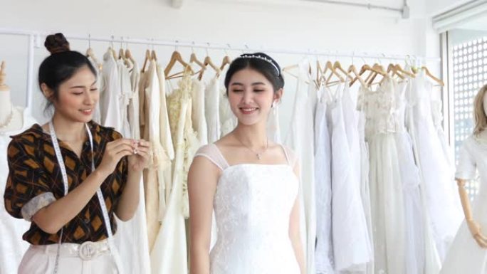 年轻美丽的亚洲新娘的小企业正站在镜子前，在服装店与时装设计师一起穿着工作室的试衣裙