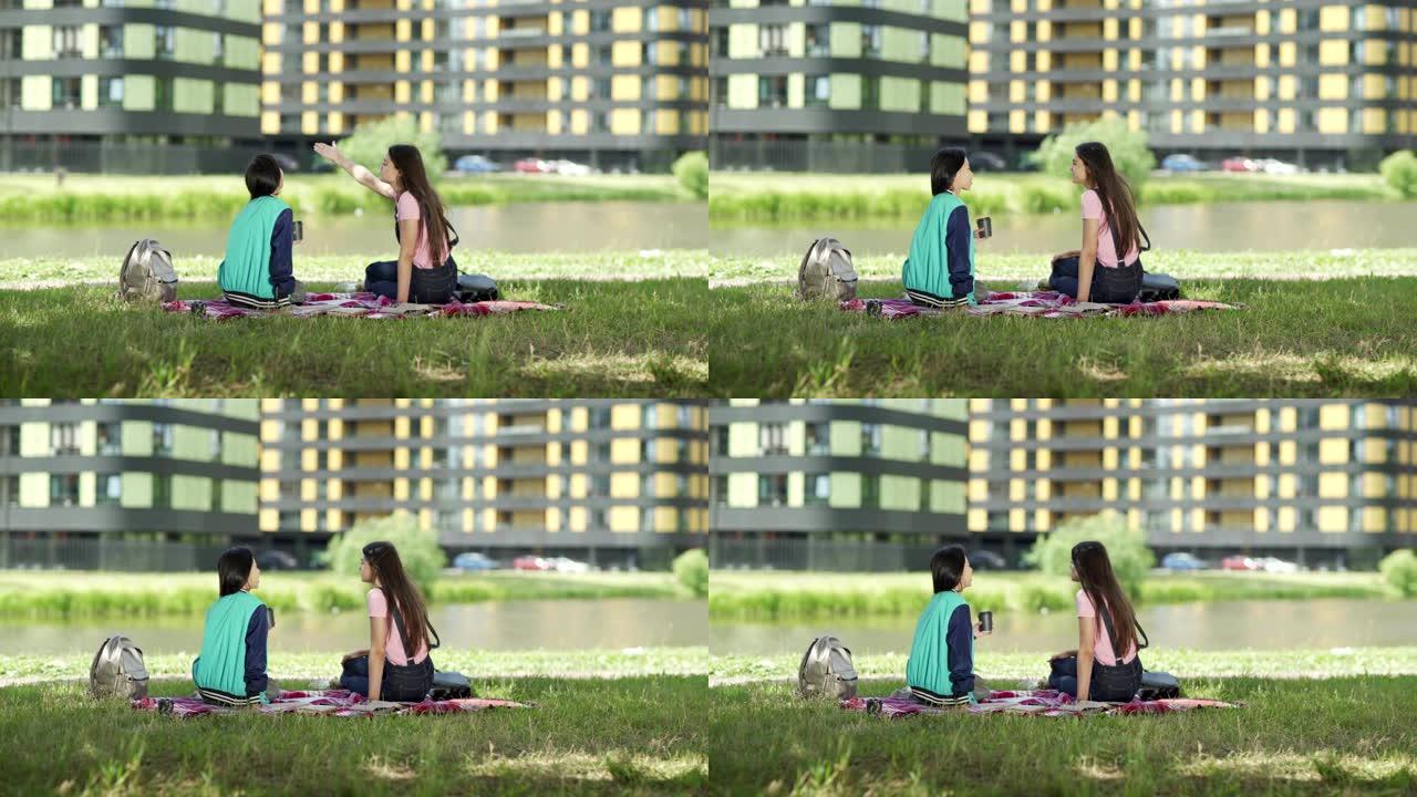 追踪两名不同的女学生坐在池塘附近的草坪上聊天后放松的镜头。亚洲女孩喝咖啡，高加索人指着校园