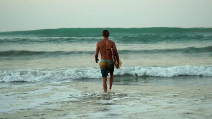 慢动作: 肌肉发达的冲浪者盖伊 (guy) 走在海滩上冲浪，在日出时拿着冲浪板。