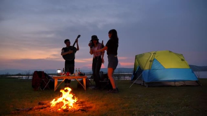 场景慢动作亚洲青少年群体在篝火帐篷前一起享受，在暑假期间在湖边观赏日落。他们在假期、放松的时刻快乐、