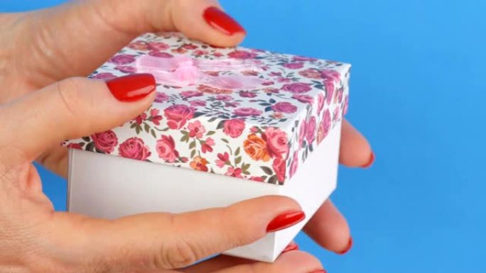 一个带有花卉图案和粉红色蝴蝶结的小礼品盒，由指甲涂有红色指甲油的女人的手握住