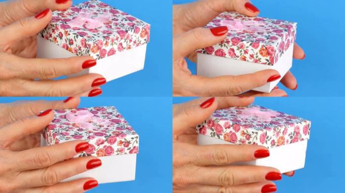 一个带有花卉图案和粉红色蝴蝶结的小礼品盒，由指甲涂有红色指甲油的女人的手握住