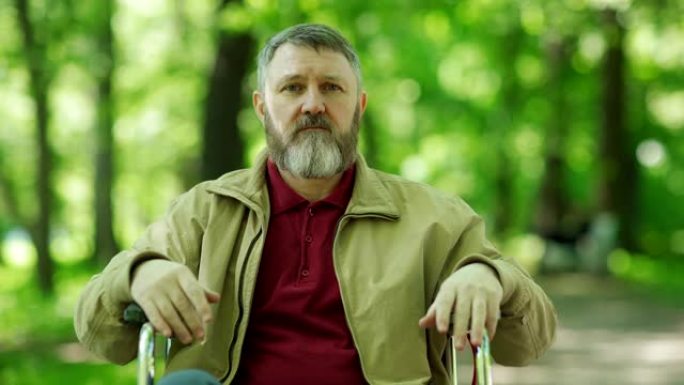 大风天绿色公园里，留着胡须的残疾成熟男人坐在轮椅上看着相机的中等镜头