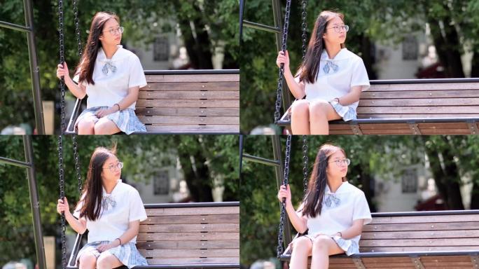 可爱的中国年轻女子，穿着日本学生风格的连衣裙，在阳光明媚的日子里，在公园的绳凳秋千上摇曳，戴着眼镜的