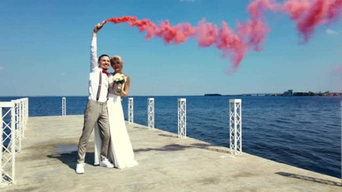 新婚夫妇的浪漫之吻，手里拿着彩色烟雾，烟雾弹。他们拥抱着，站在一个美丽的码头上，在夏天，在阳光明媚的