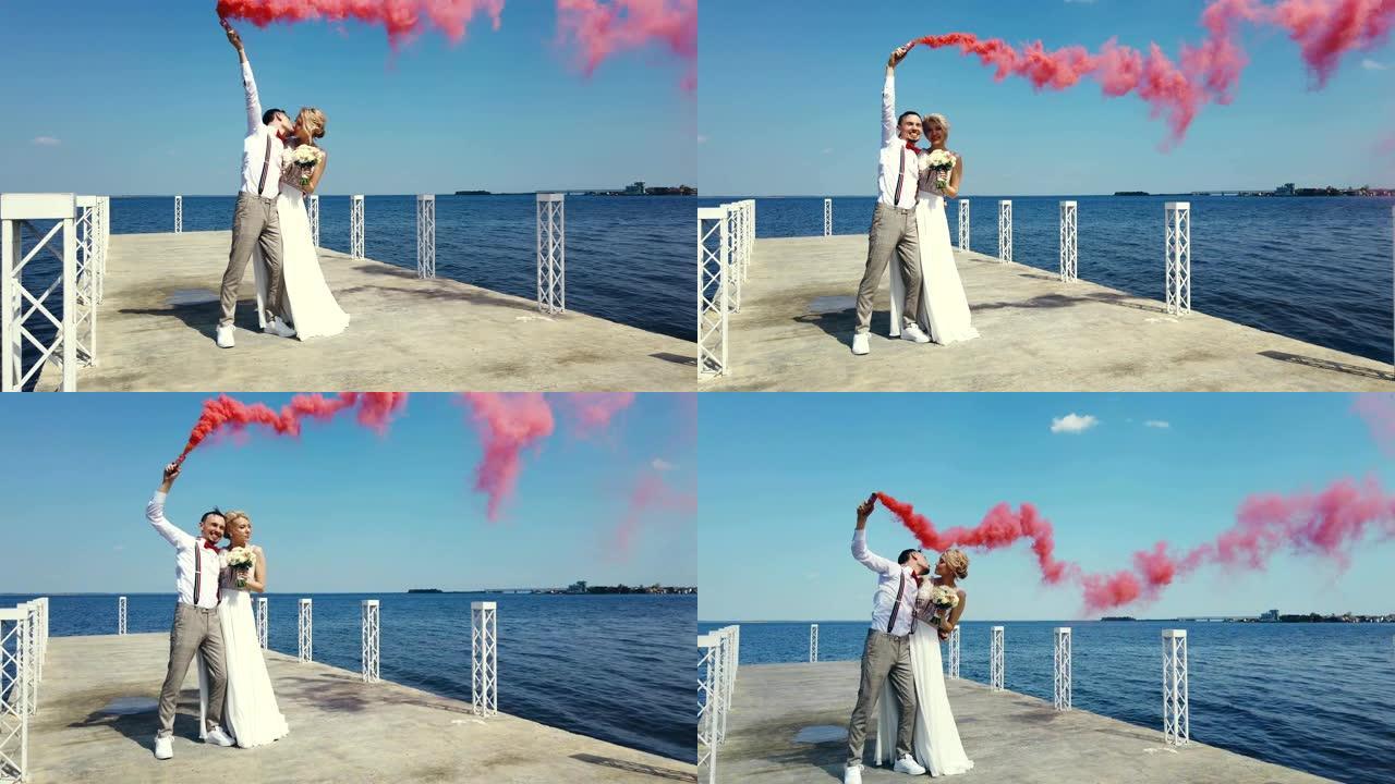 新婚夫妇的浪漫之吻，手里拿着彩色烟雾，烟雾弹。他们拥抱着，站在一个美丽的码头上，在夏天，在阳光明媚的
