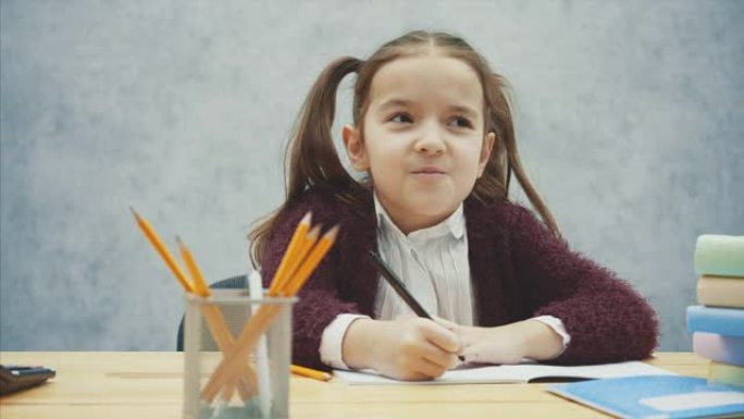 一个美丽的女学生的特写镜头坐在灰色背景的桌子旁。在此期间，他进行练习并写笔。计算习题时，取计算器。