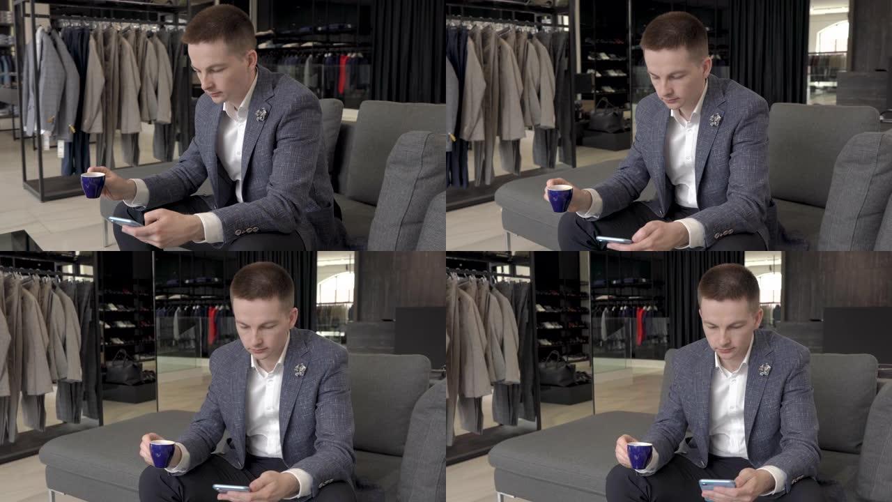 一位客户边喝咖啡边看手机屏幕。他坐在奢侈品牌男装店的现代风格灰色沙发上。旋转视频