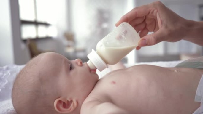 女婴从母亲手中拿着的瓶子里喝牛奶的肖像，尿布中的新生儿躺在换衣桌上特写