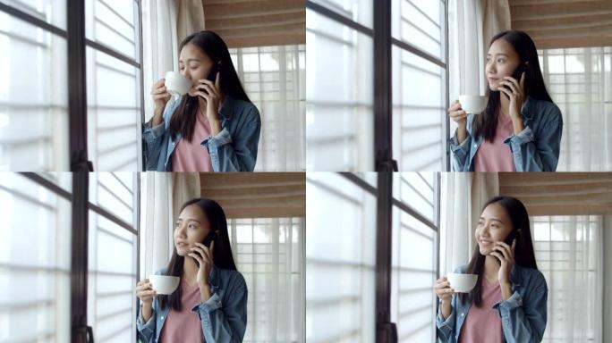 迷人的肖像微笑的年轻亚洲女性喝咖啡，和站在家庭办公室窗户旁边的朋友打电话。
