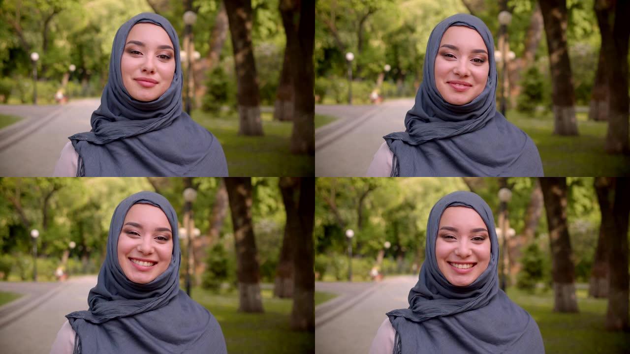 戴着头巾的穆斯林妇女的肖像，明亮的化妆害羞地微笑着，快乐地走进公园里的相机。