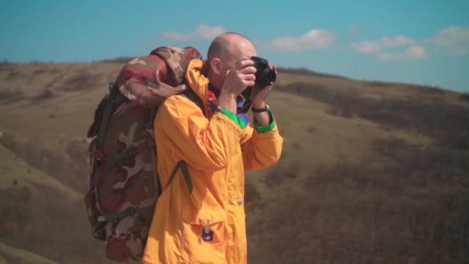 一名身穿黄色外套，戴着眼镜的男子站在山上，欣赏风景并在dslr相机上拍照。