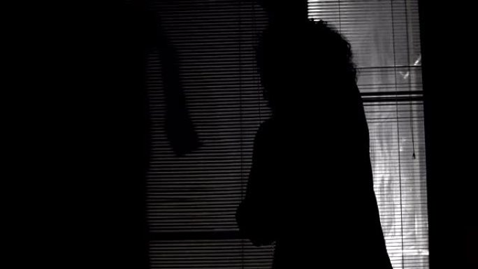孤独害怕的女人晚上关百叶窗。街灯背光女性剪影