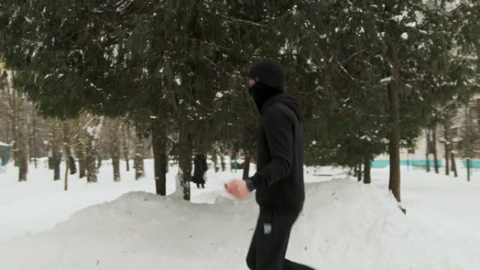年轻的家伙脸上戴着面具，穿着黑色运动服，在寒冷的天气里在户外白雪覆盖的公园里练习，跳绳
