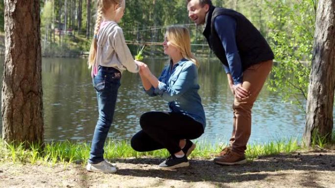 开朗的小女儿跳着父母站在乡下湖边，给幸福的母亲一小束绿草和鲜花。家庭在大自然中快乐地交谈