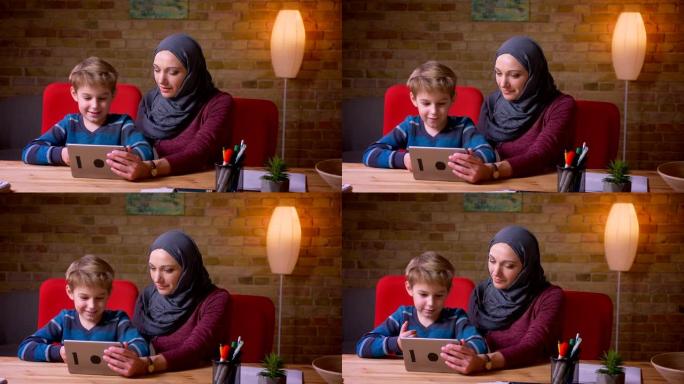 小男孩和他的穆斯林母亲戴着头巾的肖像，一起在平板电脑上观看动画片并讨论它们。