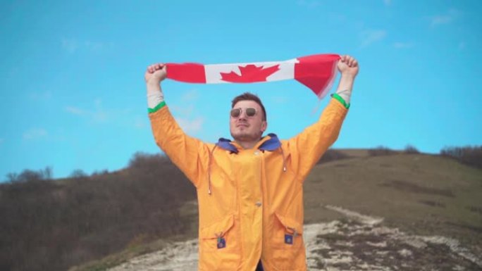 一个穿着黄色夹克、蓝色牛仔裤和眼镜的男人站在山里，双手举着加拿大国旗。