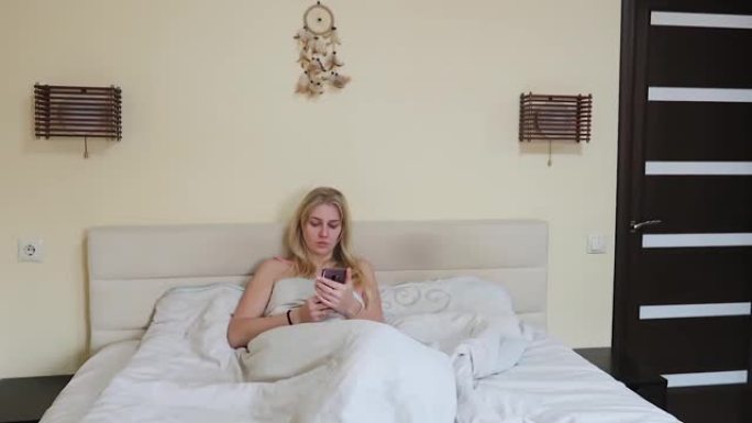 美丽的年轻女子躺在一张大床上看着手机，打着哈欠，用手捂着嘴