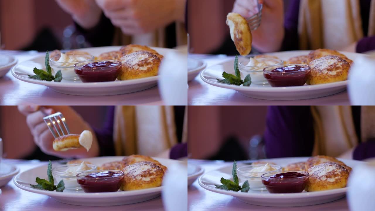 女性的手在餐厅用刀叉切了美味的奶酪煎饼。奶酪煎饼配果酱和酸奶油。