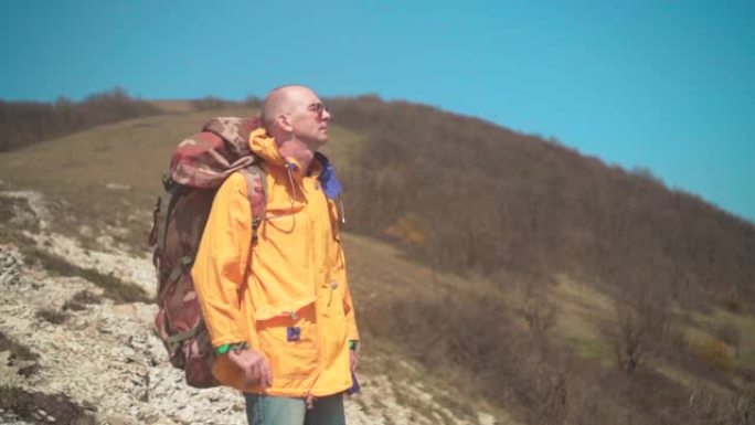 一个穿着黄色夹克、戴着眼镜、背着一个大旅游背包的男人站在山上欣赏风景。