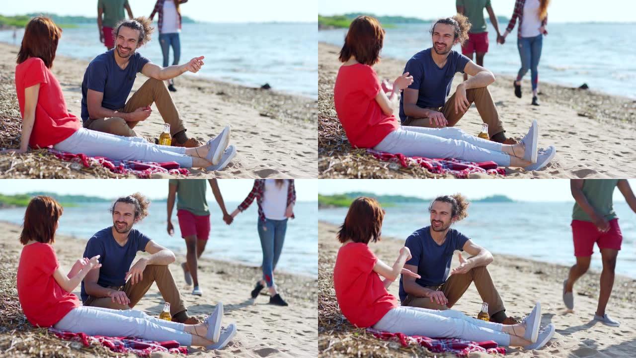 马尾辫的英俊西班牙裔男子与坐在海滩毯子上的亚洲女友交谈，另一对不同的夫妇牵着手沿着海岸线走过
