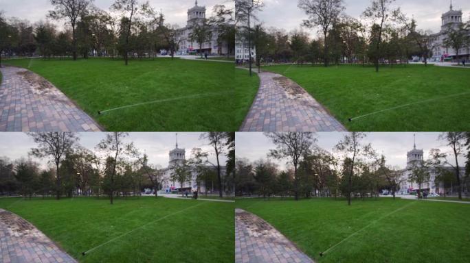 公园靠近人行道的花园灌溉洒水草坪。自动旋转灌溉系统。绿草和景观设计。滑块上的相机，4k跟踪拍摄