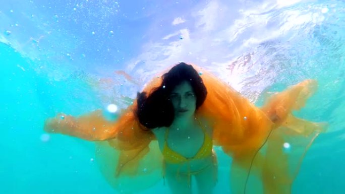一个童话般的女孩，手里拿着黄色的衣服，穿着黄色的泳衣潜入水下，在阳光明媚的日子里，她的头发朝下，向相