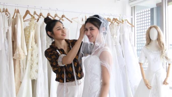 年轻美丽的亚洲新娘的小企业正站在镜子前，在服装店与时装设计师一起穿着工作室的试衣裙