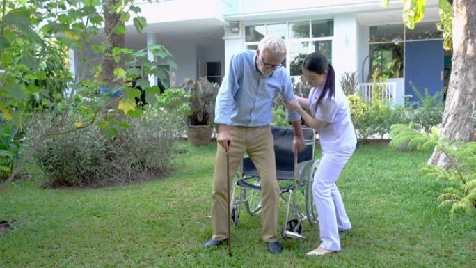 护士帮助用拐杖支持老人从养老院的花园里的轮椅上站起来-录像视频